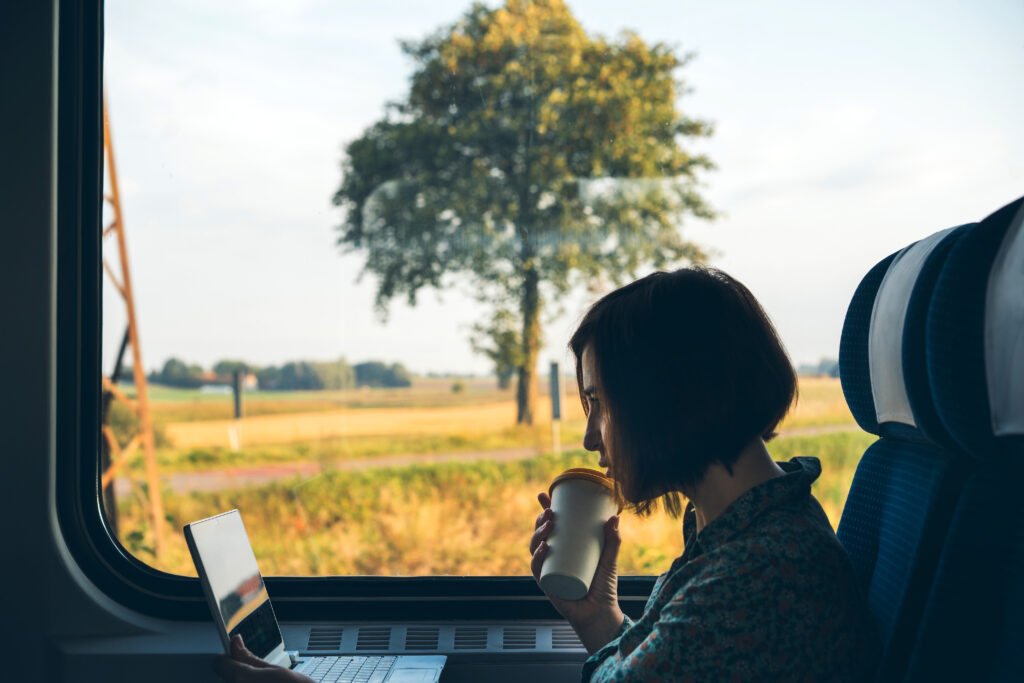 En kvinna sitter vid fönstret på ett pendeltåg med en laptop och en kaffekopp i handen.